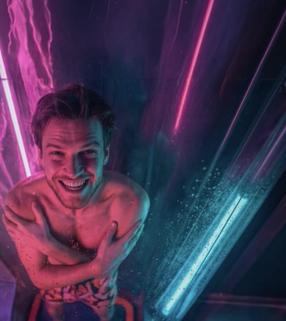A smiling man crouching down, ready to slide through a water park slide tunnel illuminated with neon pink and blue lights.