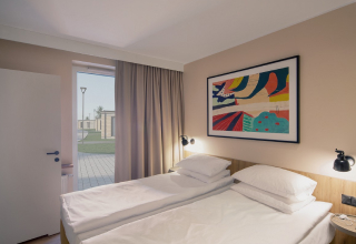 Pokój hotelowy z dwoma pojedynczymi łóżkami, nowoczesnym oświetleniem i kolorowym obrazem na ścianie; przez otwarte drzwi widać patio